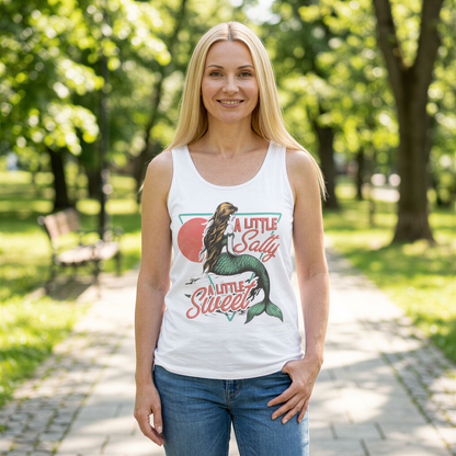 A smiling woman wearing a white tank top with a mermaid graphic and text printed on it, standing on a path in a park.