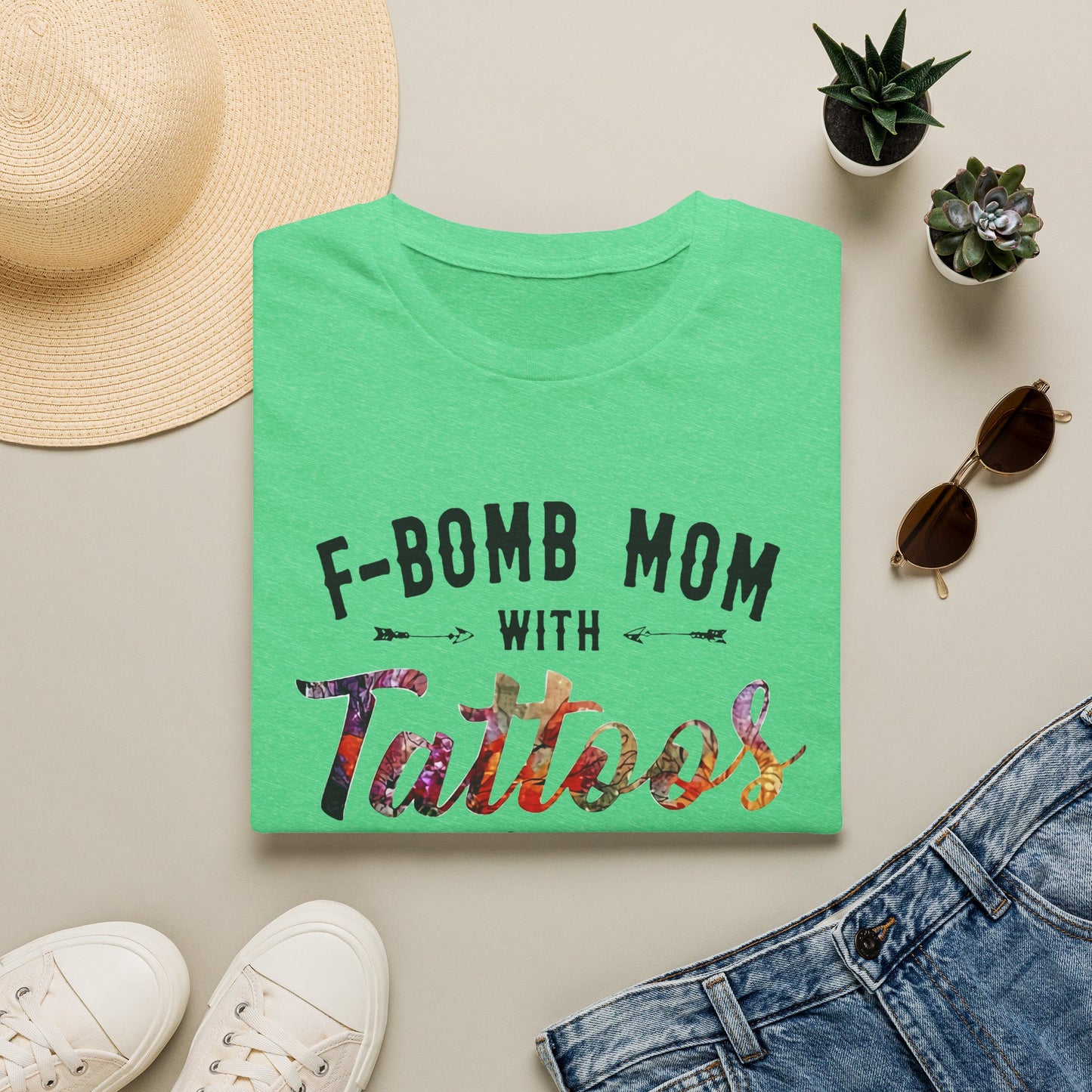 a green t-shirt with the text "F-BOMB MOM WITH Tattoos" printed on it, along with a pair of jeans, sunglasses, and a straw hat.