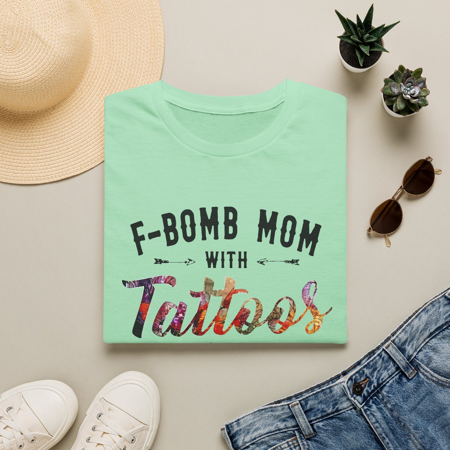 a green t-shirt with the text "F-BOMB MOM WITH TATToos" printed on it, along with a pair of jeans, sunglasses, and a straw hat.