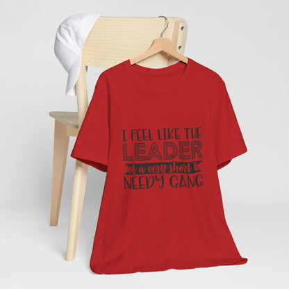 A red t-shirt with the text "I feel like the leader of a very short & NEEDY GANG" printed on it, along with a wooden chair and a white hat in the background.