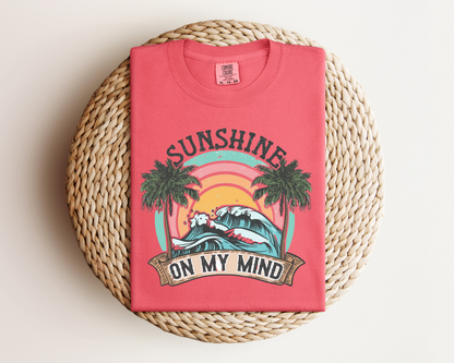A red t-shirt with a graphic design depicting a sunset, palm trees, and the text "SUNSHINE ON MY MIND" printed on it.