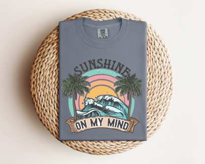 A gray t-shirt with a graphic design depicting a sunset, palm trees, and the text "SUNSHINE ON MY MIND" printed on it.