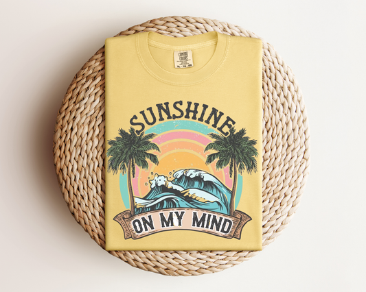 A yellow t-shirt with a graphic design featuring a palm tree, a sunset, and the text "SUNSHINE ON MY MIND" printed on it.
