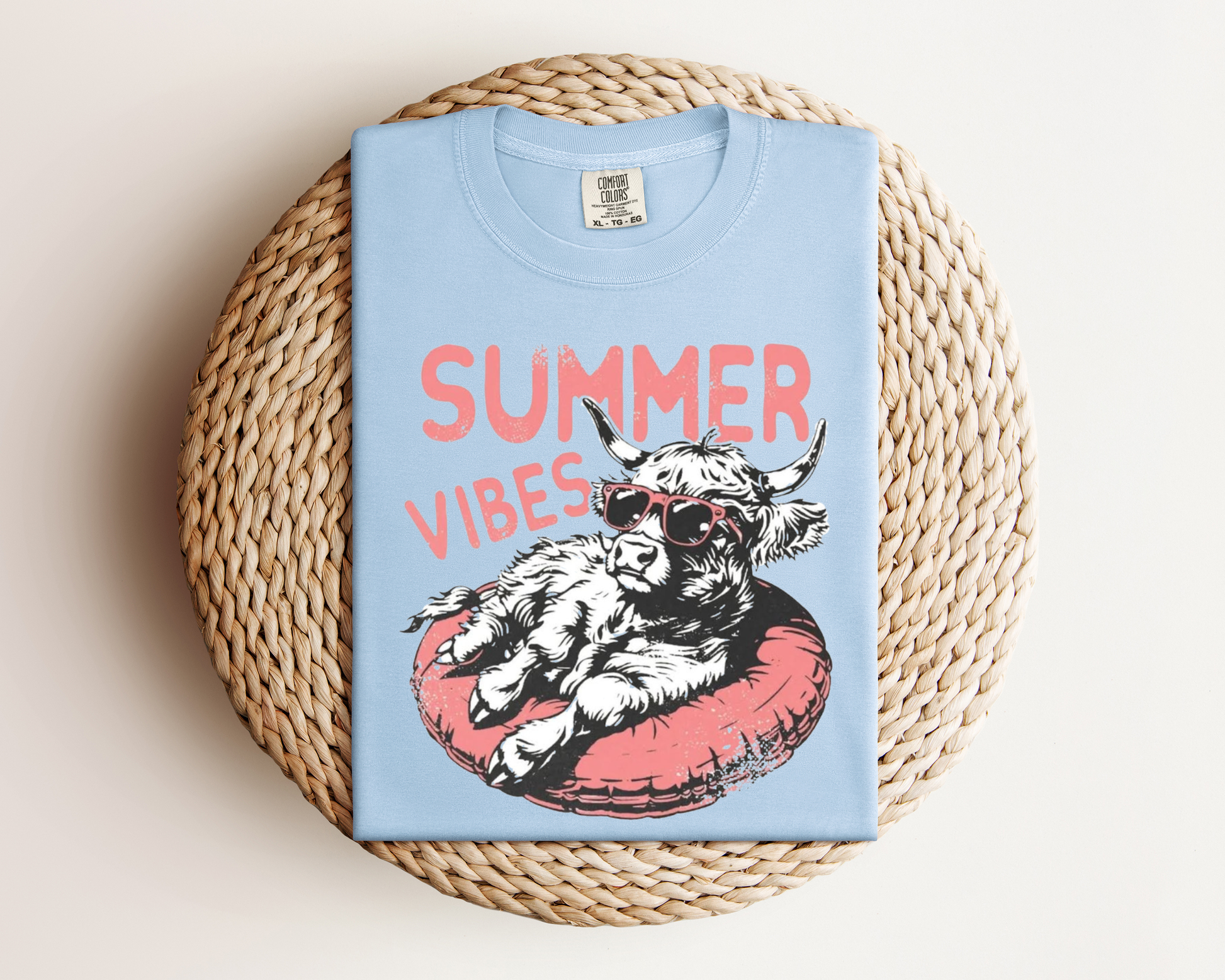 A light blue t-shirt with a graphic design of a cow wearing sunglasses and resting on a red inner tube, with the text "SUMMER VIBES" printed on it.
