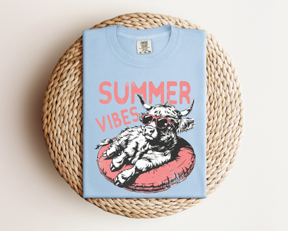 A light blue t-shirt with a graphic design of a cow wearing sunglasses and resting on a red inner tube, with the text "SUMMER VIBES" printed on it.